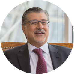 Adnan Amin busy smiling while wearing agray suit, glasses and a maroon tie
